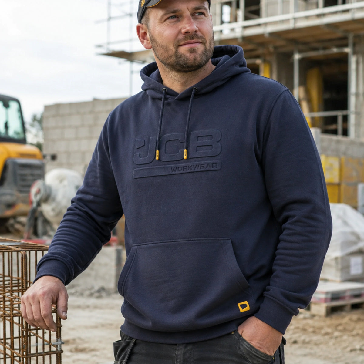 Mężczyzna w granatowej bluzie JCB z kapturem na placu budowy, ujęcie z przodu